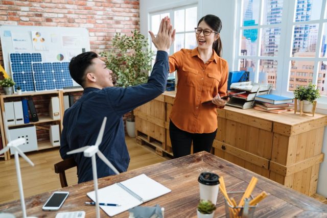 7 tips to reduce business electricity costs. Positive nice team of woman and man engineers successful saving energy in wooden office. male and female coworkers cheerful in modern workplace giving high five and celebrating done project in city